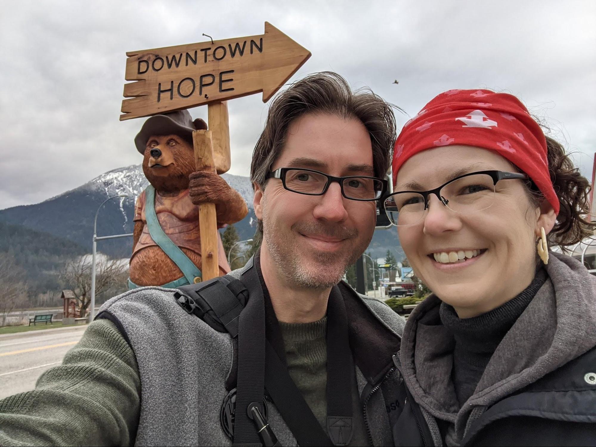 woman and man standing in front of a chainsaw carving of a bear wearing blue overalls, a brown hat, and holding a directional sign pointing to downtown Hope. 