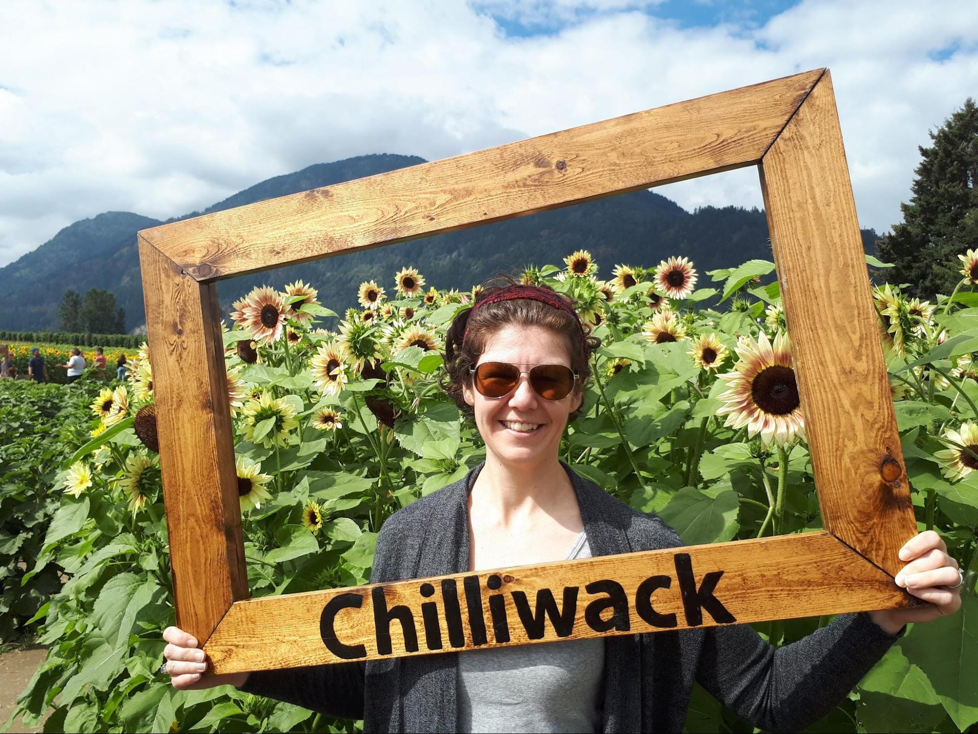 Mel holding a large frame that says "Chilliwack" and standing in front of a field of sunflowers at the Chilliwack sunflower festival.