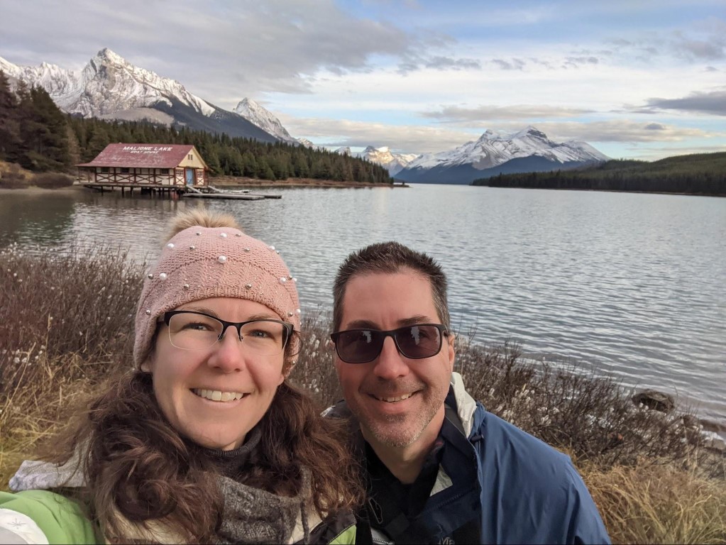 Mel and Jay standing lakeside with the Maligne Lake boathouse, Maligne Lake, and mountains in the background 