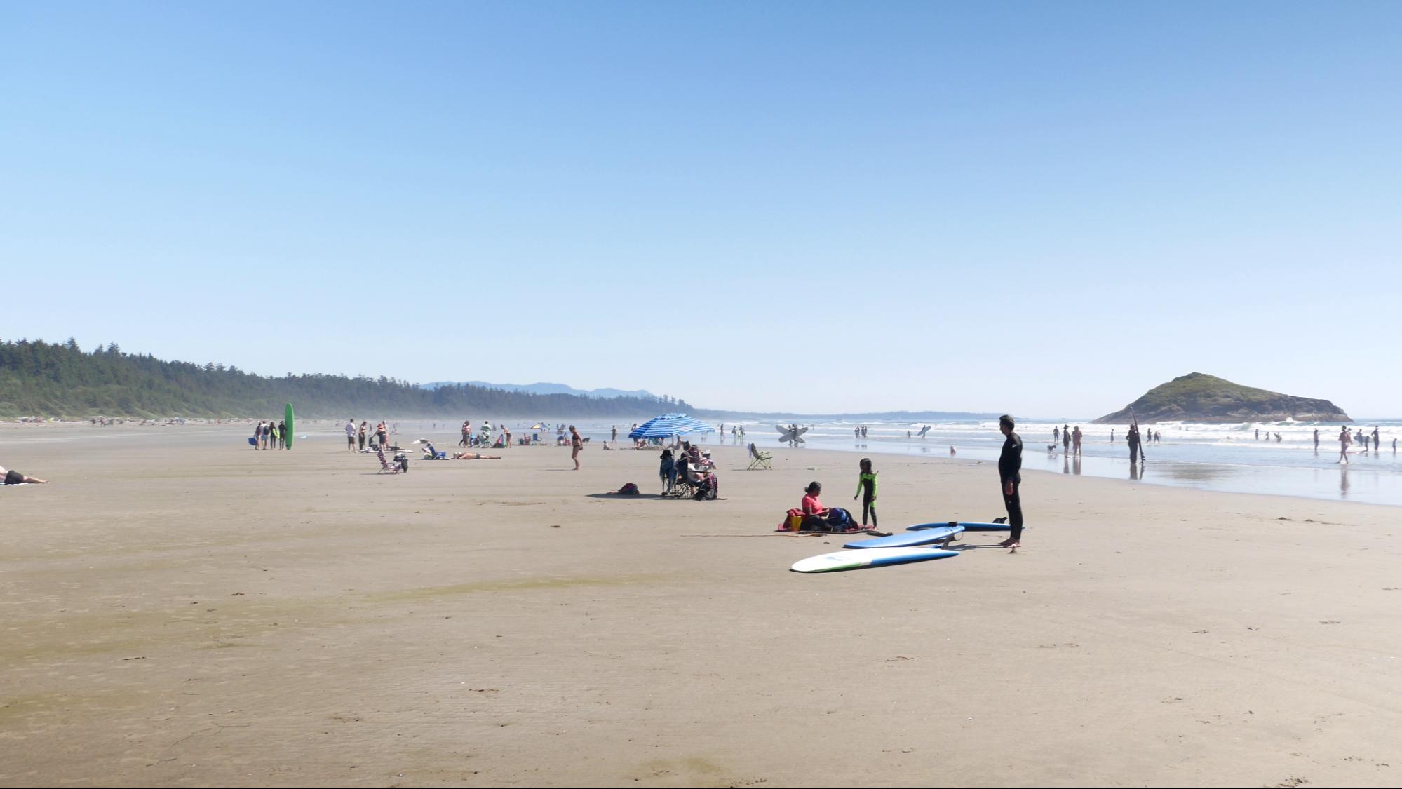 Long beach on a sunny day in Tofino. Lots of people enjoying the water, waves, and sun. 